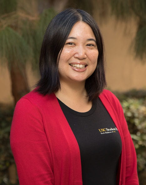 Portrait style photo of Jennifer Hsia, who is photographed standing outside in front of a wall that is covered with bushes and a tree. She is wearing a black jacket and black t-shirt that says "USC Student" Health.